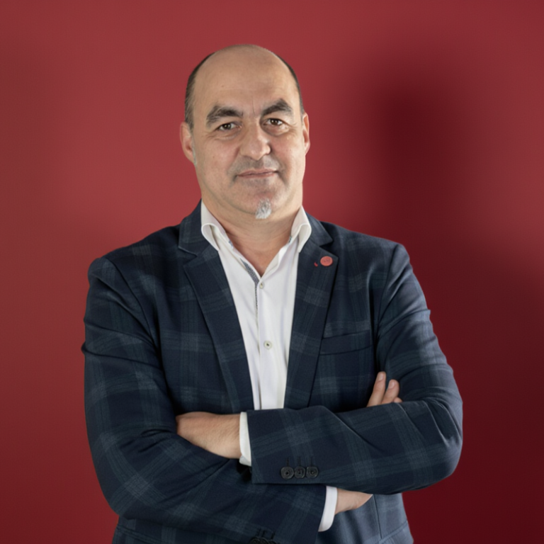 tu gestor personal Hombre de pie con brazos cruzados, sonriente, vestido con chaqueta sobre fondo rojo.